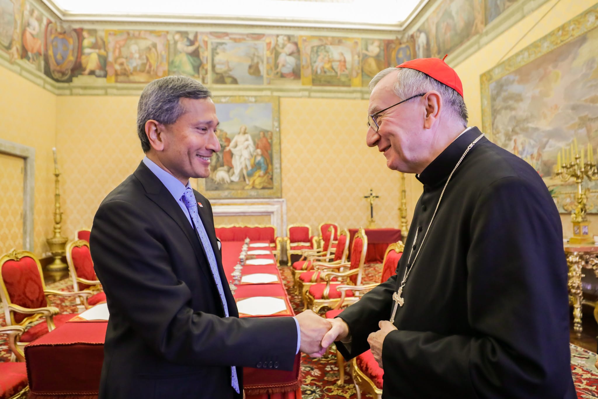 Two men shaking hands in ornate room, one in suit, the other in cardinal's garb.